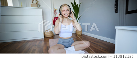 Young and sporty woman working out at home, doing fitness training alone on yoga mat, listens to music in wireless headphones Young and sporty woman working out at home, doing fitness training alone on yoga mat, listens to music in wireless headphones 113812185