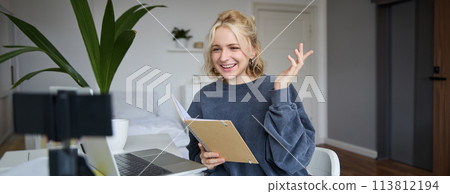 Portrait of young smiling blond woman, working from home, online chatting, using digital video camera, recording vlog, holding notebook, reading notes, explaining something 113812194
