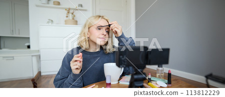 Portrait of beautiful, smiling blond woman, girl recording video of her makeup tutorial for social media, vlogger sitting on floor in her room, using stabiliser to create content, reviewing mascara Portrait of beautiful, smiling blond woman, girl recording video of her makeup tutorial for social media, vlogger sitting on floor in her room, using stabiliser to create content, reviewing mascara 113812229
