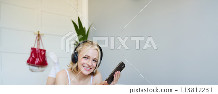 Vertical shot of young fitness woman in headphones, lying on rubber mat, relaxing after workout training session, holding smartphone, smiling at camera 113812231