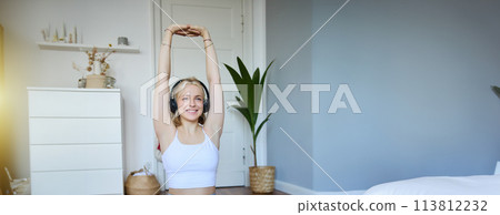 Portrait of woman doing fitness exercises at home, watches sports video on laptop, wearing wireless headphones, follows workout instructions, sits on rubber mat in her room 113812232