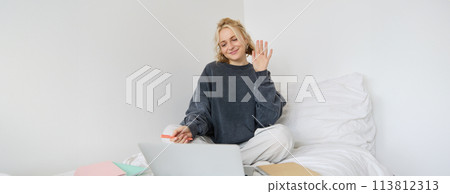 Portrait of beautiful, smiling young woman sitting on bed, waves her hand at laptop camera, says hello to friend on video chat app, studies at home on remote, connects to online course 113812313