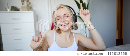 Portrait of happy and carefree blond woman, listens to music in wireless headphones, dancing and singing along favourite song, spending time alone at home 113812366