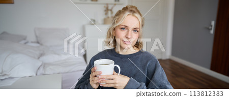 Close up portrait of smiling young blond woman, sits with cup of tea in bedroom, rests at home, enjoys her coffee Close up portrait of smiling young blond woman, sits with cup of tea in bedroom, rests at home, enjoys her coffee 113812383