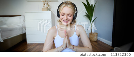 Image of young woman listens to yoga podcast for beginners, holds hands in namaste sign, wears headphones, meditates 113812390