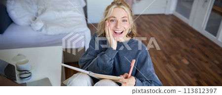 Portrait of happy, young blond woman, sitting in bedroom with notebook and pen, laughing and smiling, writing in journal, doing homework and smiling 113812397