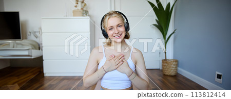 Portrait of young happy woman in headphones, looking with relaxed, friendly face expression, holding hands on her chest and smiling Portrait of young happy woman in headphones, looking with relaxed, friendly face expression, holding hands on her chest and smiling 113812414