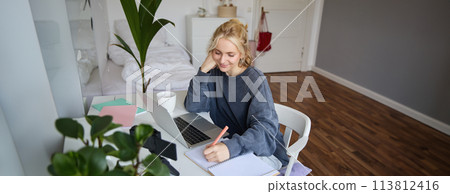 Lifestyle image of young woman writing down something in notebook, making notes, studying online, doing course in internet, listening to interesting information, using laptop 113812416