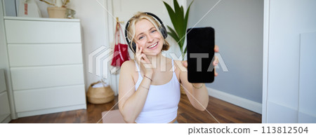 Portrait of smiling, cheerful young woman workout at home and showing smartphone screen, wearing headphones, recommending mobile app 113812504