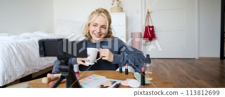Blogging, modern technology, e-commerce and people concept. Happy smiling woman beauty video blogger holding cup of tea applying makeup while recording beauty tutorial at home for her video blog 113812699