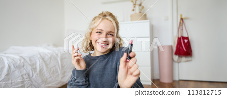Portrait of young social media influencer, woman recording a video with beauty products, showing her makeup on camera, holding lipstick and smiling, sitting on floor Portrait of young social media influencer, woman recording a video with beauty products, showing her makeup on camera, holding lipstick and smiling, sitting on floor 113812715