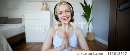 Portrait of young relaxed woman, sits in room in headphones, clasp hands together, meditated, listens to yoga music 113812717
