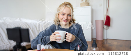 Close up portrait of beautiful blond woman, girl sits in her room, does makeup, records video for social media, drinks tea, holds mug and smiles at camera 113812718