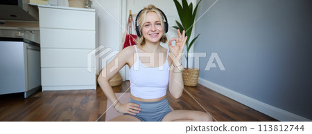Portrait of cute smiling blond woman wearing wireless headphones, workout at home on rubber yoga mat, sitting in a room, showing okay, yes hand sign Portrait of cute smiling blond woman wearing wireless headphones, workout at home on rubber yoga mat, sitting in a room, showing okay, yes hand sign 113812744