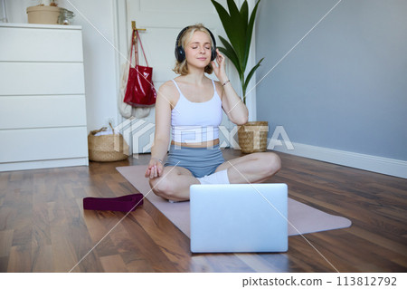 Portrait of fit and healthy woman at home, practice yoga, sitting on rubber mat, listening to instructions online, using meditation music to relax, following guidance on laptop 113812792