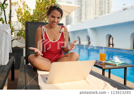 Smiling woman in red swimsuit video calling coworker when sunbathing by pool Smiling woman in red swimsuit video calling coworker when sunbathing by pool 113813293
