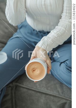A woman sits on the couch holding jar with cosmetic cream A woman sits on the couch holding jar with cosmetic cream 113814022