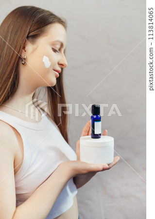 Woman with cream on face skin holding daily set of essential cosmetics jar of cream and serum bottle 113814065