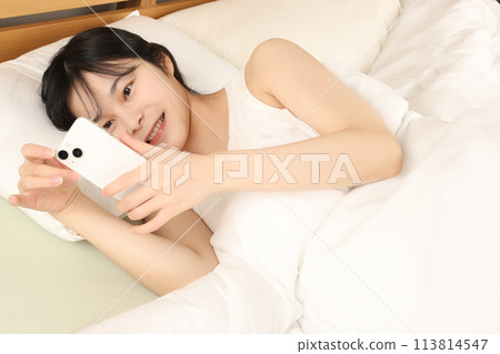 Smiling young woman looking at her smartphone while lying in bed 113814547