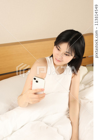 Young woman sitting in bed and looking at her smartphone Young woman sitting in bed and looking at her smartphone 113814549