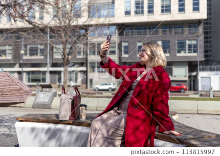 Urban Selfie Serenity: plus size blonde beauty in red coat and glasses. Holding smartphone and smiling at camera. Blogging Urban Selfie Serenity: plus size blonde beauty in red coat and glasses. Holding smartphone and smiling at camera. Blogging 113815997