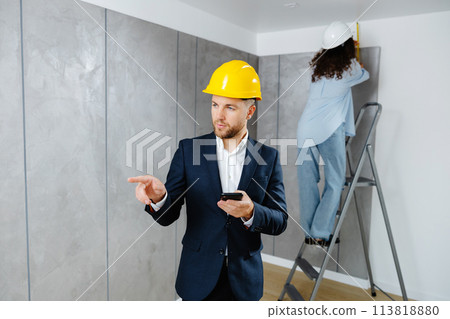 A young businessman holds a phone in his hands and checks the work of a designer in creating an interior in a room. The concept of repair on a modern design. 113818880