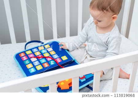 Baby sitting in a crib playing with montessori busy book. Concept of keeping children from screen by activity books and quiet books 113819068
