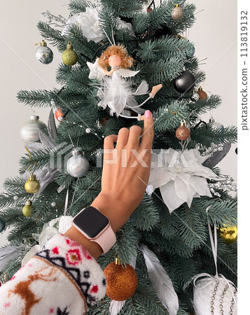Female hand with smart watch touching white little angel on Christmas tree. Female hand with smart watch touching white little angel on Christmas tree. 113819132