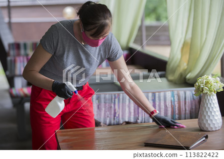 Waiter wearing protective face mask while disinfecting tables at outdoor cafe 113822422
