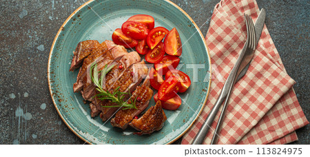 Delicious roasted sliced duck breast fillet with golden crispy skin, with pepper and rosemary, top view on ceramic blue plate served with cherry tomatoes salad, rustic concrete rustic background Delicious roasted sliced duck breast fillet with golden crispy skin, with pepper and rosemary, top view on ceramic blue plate served with cherry tomatoes salad, rustic concrete rustic background 113824975