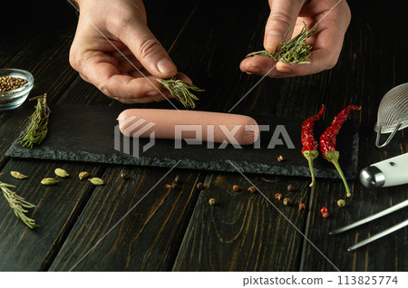 Cooking Munich sausage. The chef hands add fragrant rosemary to the sausage on the kitchen table before baking in the kitchen. Tasty snack serving concept Cooking Munich sausage. The chef hands add fragrant rosemary to the sausage on the kitchen table before baking in the kitchen. Tasty snack serving concept 113825774