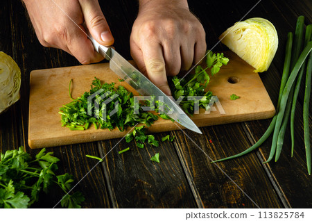 The cook is preparing a salad on the kitchen table. Close-up of a chef hands using a knife to chop fresh green parsley The cook is preparing a salad on the kitchen table. Close-up of a chef hands using a knife to chop fresh green parsley 113825784