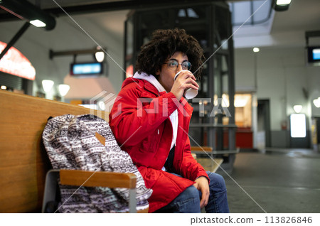 Young curly-haired guy having coffee and waiting for the train at the railway station Young curly-haired guy having coffee and waiting for the train at the railway station 113826846