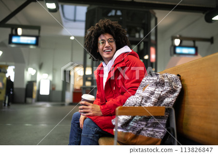 Young curly-haired guy having coffee and waiting for the train at the railway station Young curly-haired guy having coffee and waiting for the train at the railway station 113826847