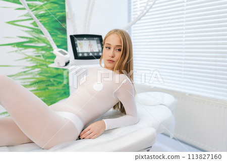 Medium shot of blonde female patient in white uniform lying on medical couch in modern health center while waiting beautician specialist. Concept of cosmetology, spa, beauty treatment. Medium shot of blonde female patient in white uniform lying on medical couch in modern health center while waiting beautician specialist. Concept of cosmetology, spa, beauty treatment. 113827160