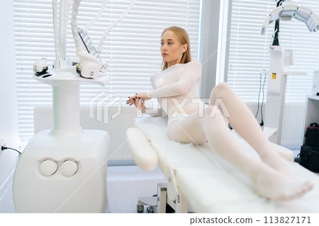 Portrait of attractive blonde female patient in white uniform lying on medical couch in modern health center while waiting beautician specialist. Concept of cosmetology, spa, beauty treatment. 113827171