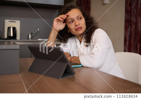Emotional portrait of a stressed middle aged woman dressed in white bathrobe at home, looking away, tired from learning on digital tablet. Emotional stress. Sadness. Depression. Frustration. Burnout. 113828634