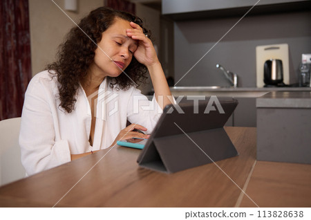 Tired business woman in white bathrobe at home, working online on tablet, reading email with bad news, suffering from burnout, sitting at kitchen table. Young female boss dealing with work problem 113828638