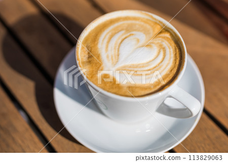 Coffee on wooden background. Cup of cappuccino with latte art on brown table with sunbeam. Fresh morning coffee with delicious milk foam. Coffee on wooden background. Cup of cappuccino with latte art on brown table with sunbeam. Fresh morning coffee with delicious milk foam. 113829063
