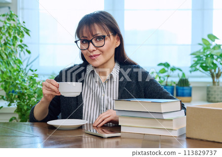 Middle aged woman holding cup of coffee, sitting at table with pile of books Middle aged woman holding cup of coffee, sitting at table with pile of books 113829737