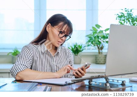 Middle aged woman working with computer, using smartphone, in home office Middle aged woman working with computer, using smartphone, in home office 113829739