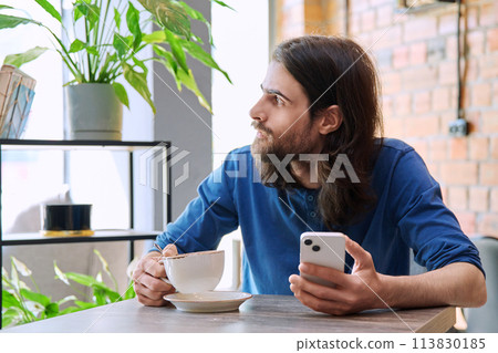 Young handsome man using smartphone, drinking cup of coffee, sitting in cafe Young handsome man using smartphone, drinking cup of coffee, sitting in cafe 113830185