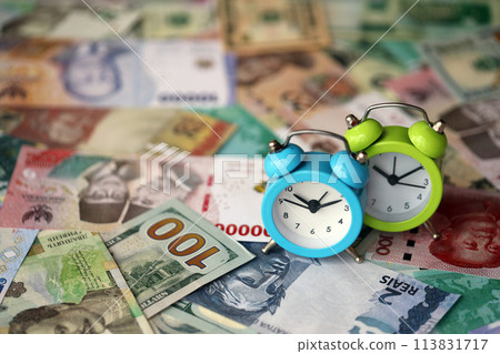 Small alarm clock on many banknotes of different currency. Background of time and money close up Small alarm clock on many banknotes of different currency. Background of time and money close up 113831717