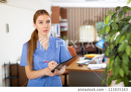 Young woman therapist writes notes on a tablet, standing in a office Young woman therapist writes notes on a tablet, standing in a office 113835163