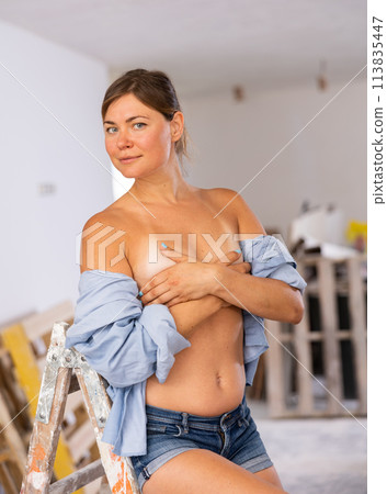 Seminude woman in unbuttoned shirt covering breasts with hands in room during renovation 113835447