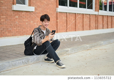 A positive Asian male college student sits on the street near the campus building using his phone. A positive Asian male college student sits on the street near the campus building using his phone. 113835722