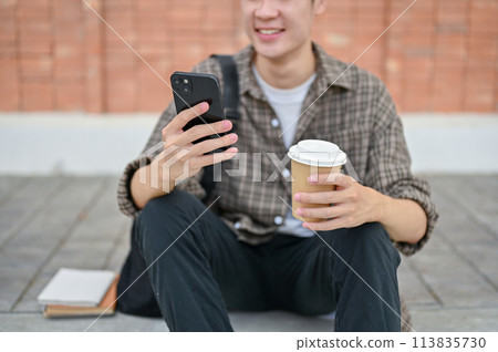 A young male college student sits on the street using his smartphone and holding a coffee cup. 113835730