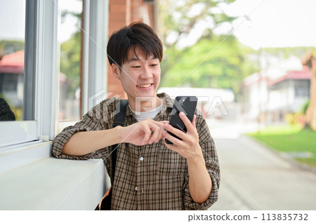 A smiling young Asian male college student stands by the campus building using his smartphone. 113835732
