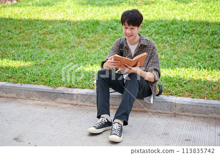 A positive Asian male college student sits on the street in the campus park reading a book. 113835742