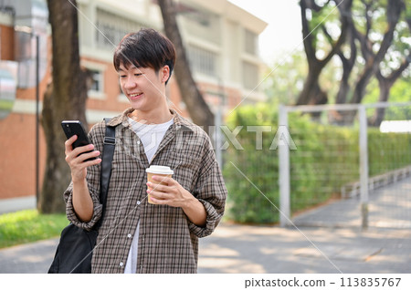 A handsome, positive young Asian man in casual wear using his smartphone while walking in a park. 113835767